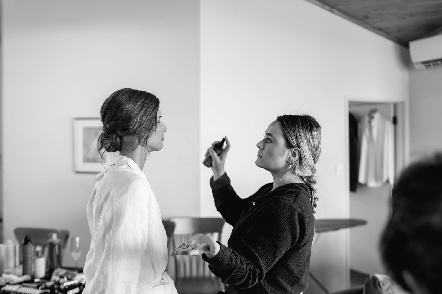 Black and white photo of hairstylist applying makeup in bright indoor setting.