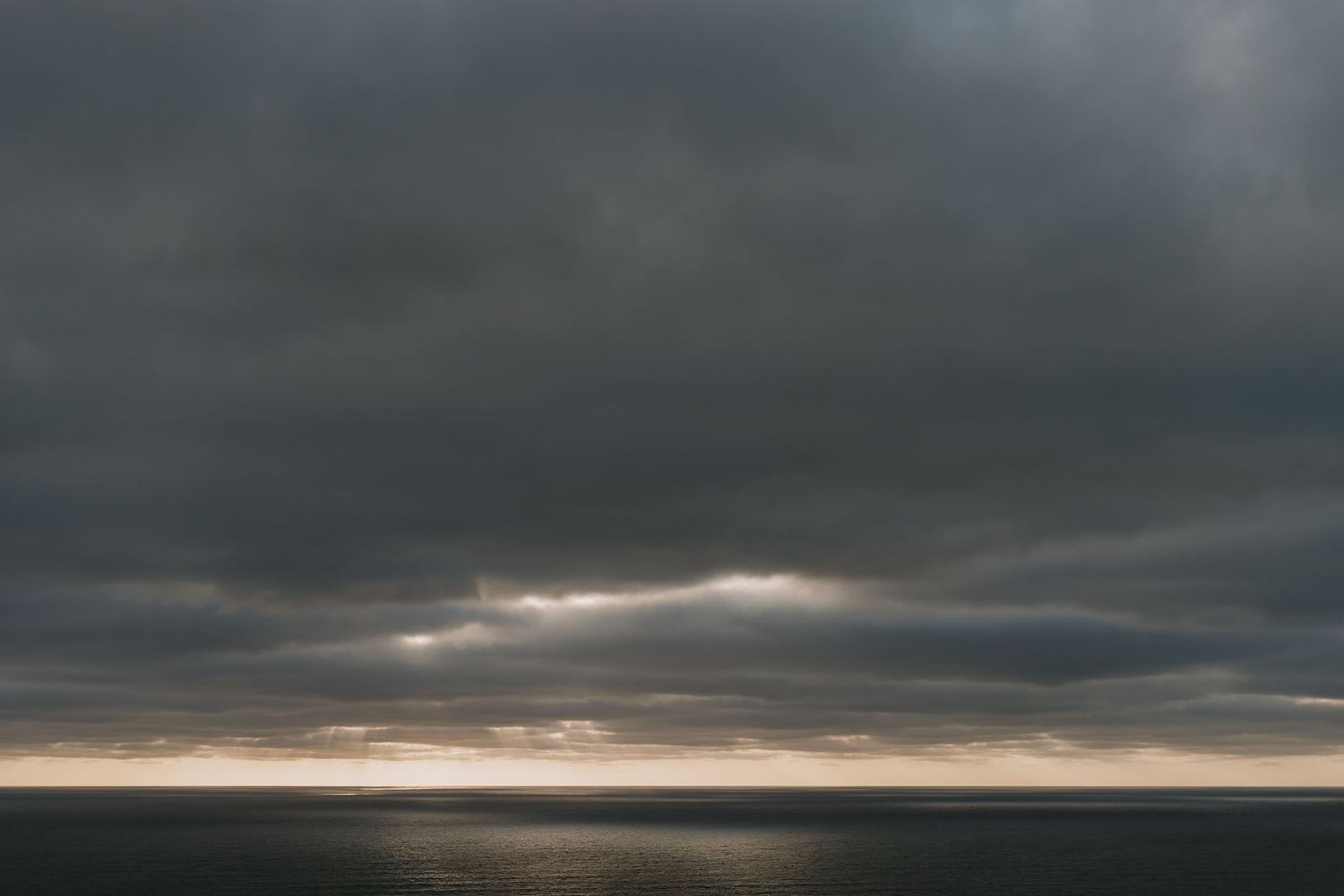 Dark storm clouds loom over a moody seascape with sunlight breaking through on the horizon.