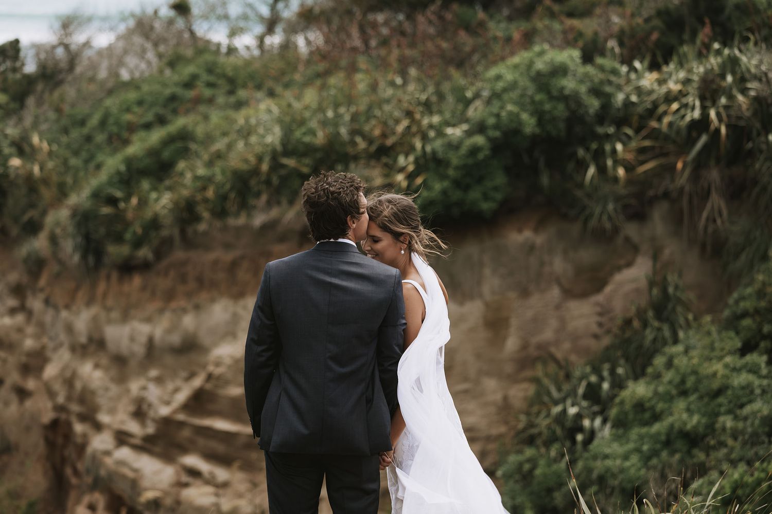 Newlywed couple stands on cliff edge overlooking natural scenery in elegant wedding attire.