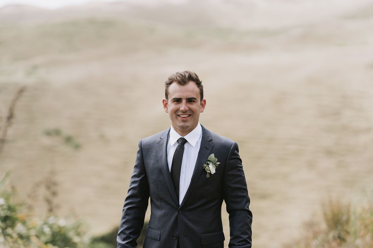 A group of formally dressed groomsmen pose together on a grassy hillside during a wedding photo session.