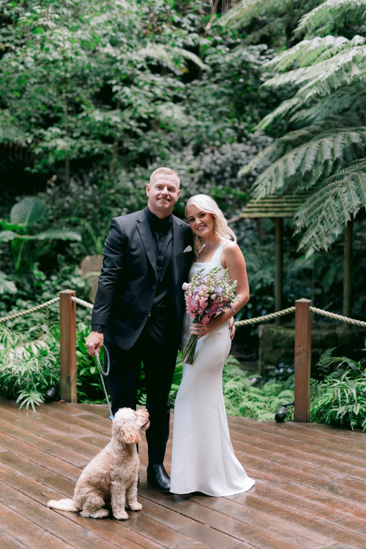 Elegant wedding couple poses with their goldendoodle dog on a wooden deck surrounded by lush tropical greenery.