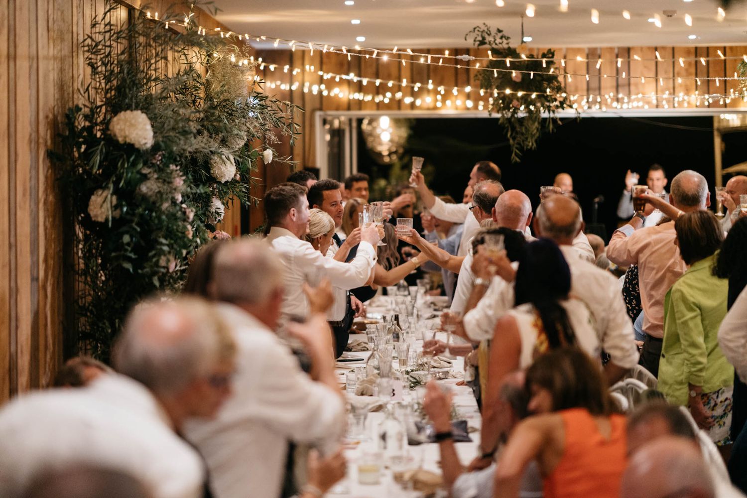 Wedding reception guests dance and celebrate under warm string lights in an elegant indoor venue setting.