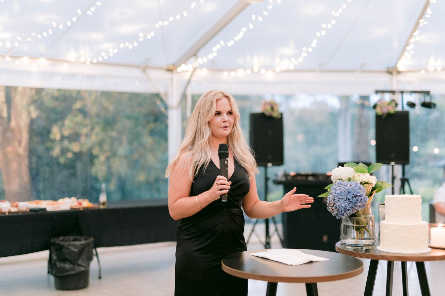Wedding event speaker in a black dress gestures on stage under an elegant white tent with floral decorations.
