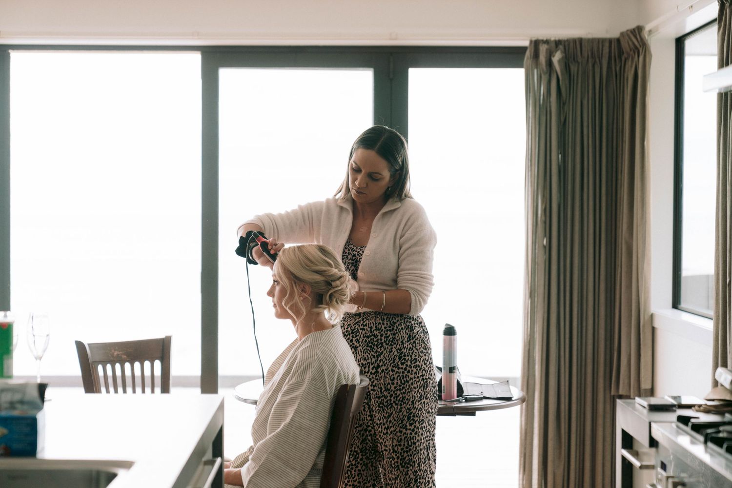 A hairstylist wearing a white blouse and patterned skirt styles someone's hair in a bright, window-lit room.