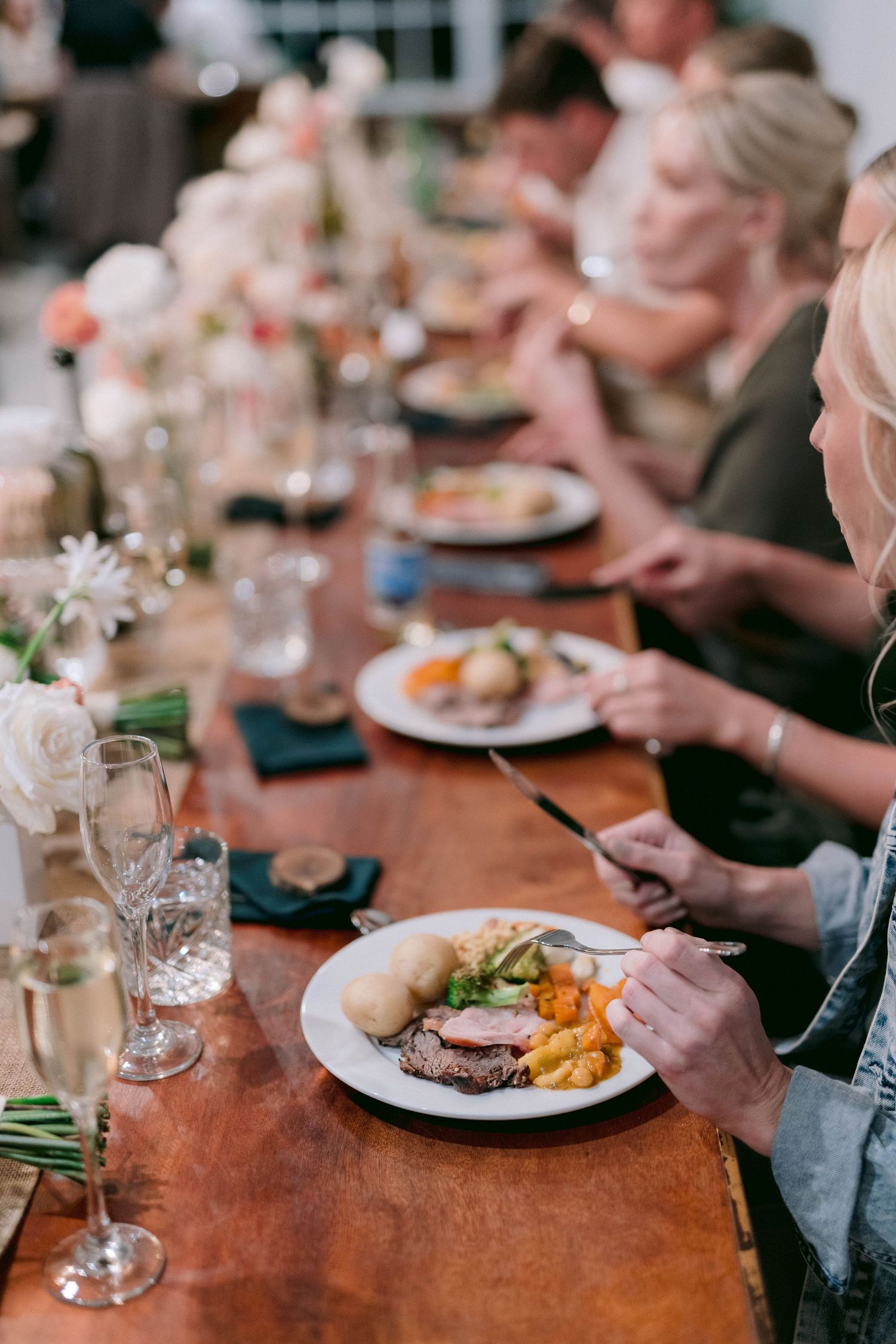 Wedding guests dining at a long wooden table with elegant place settings and floral centerpieces.