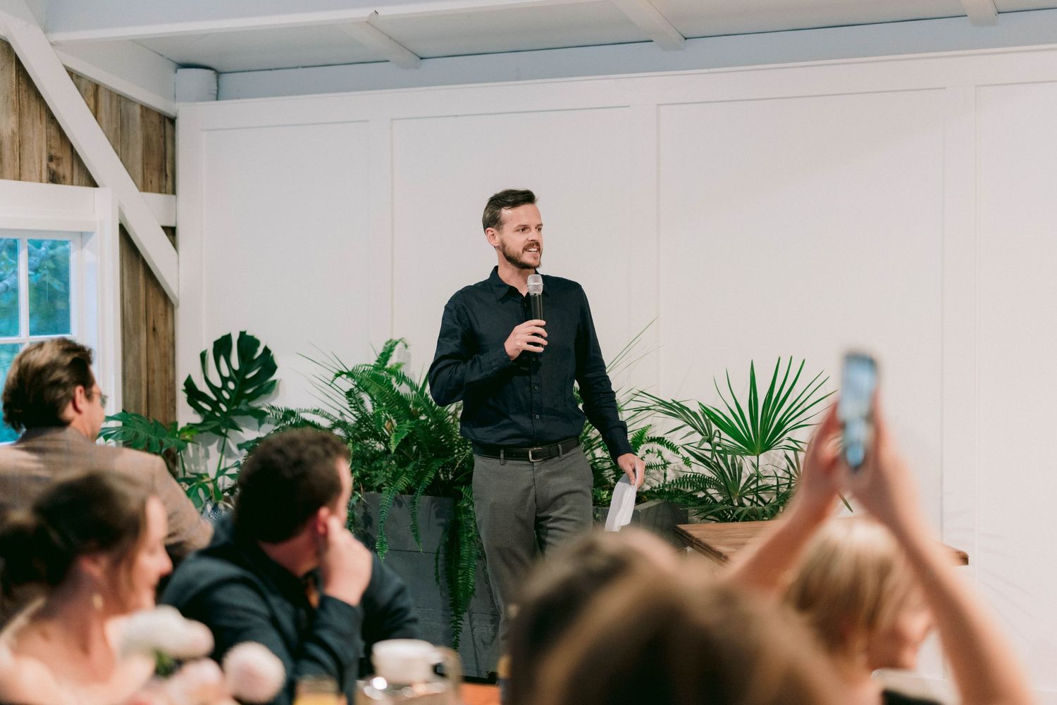 Speaker addressing wedding guests in modern white room with tropical plants.