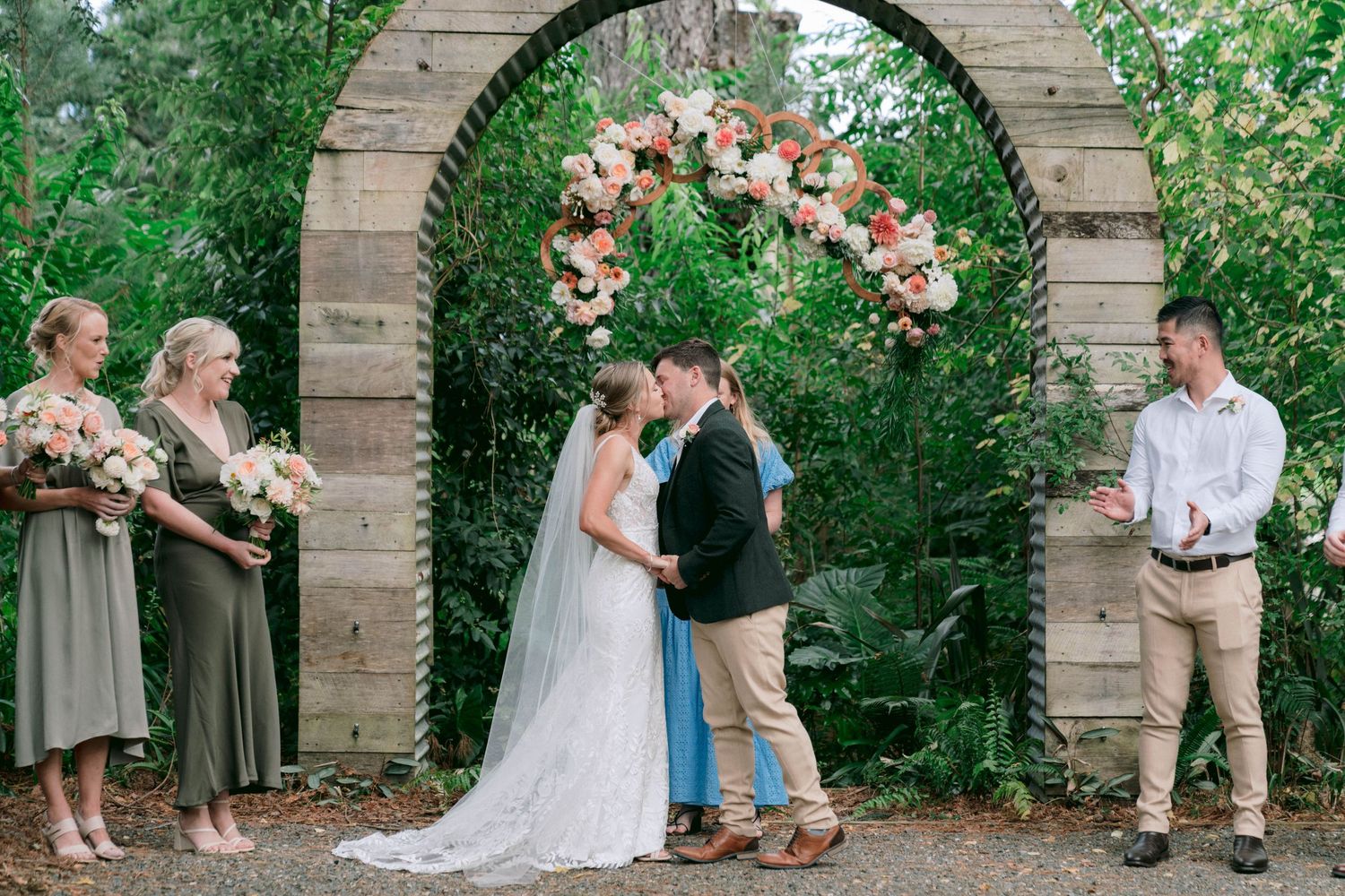 Wedding ceremony under a brick archway decorated with pink and peach floral arrangements in a garden setting.