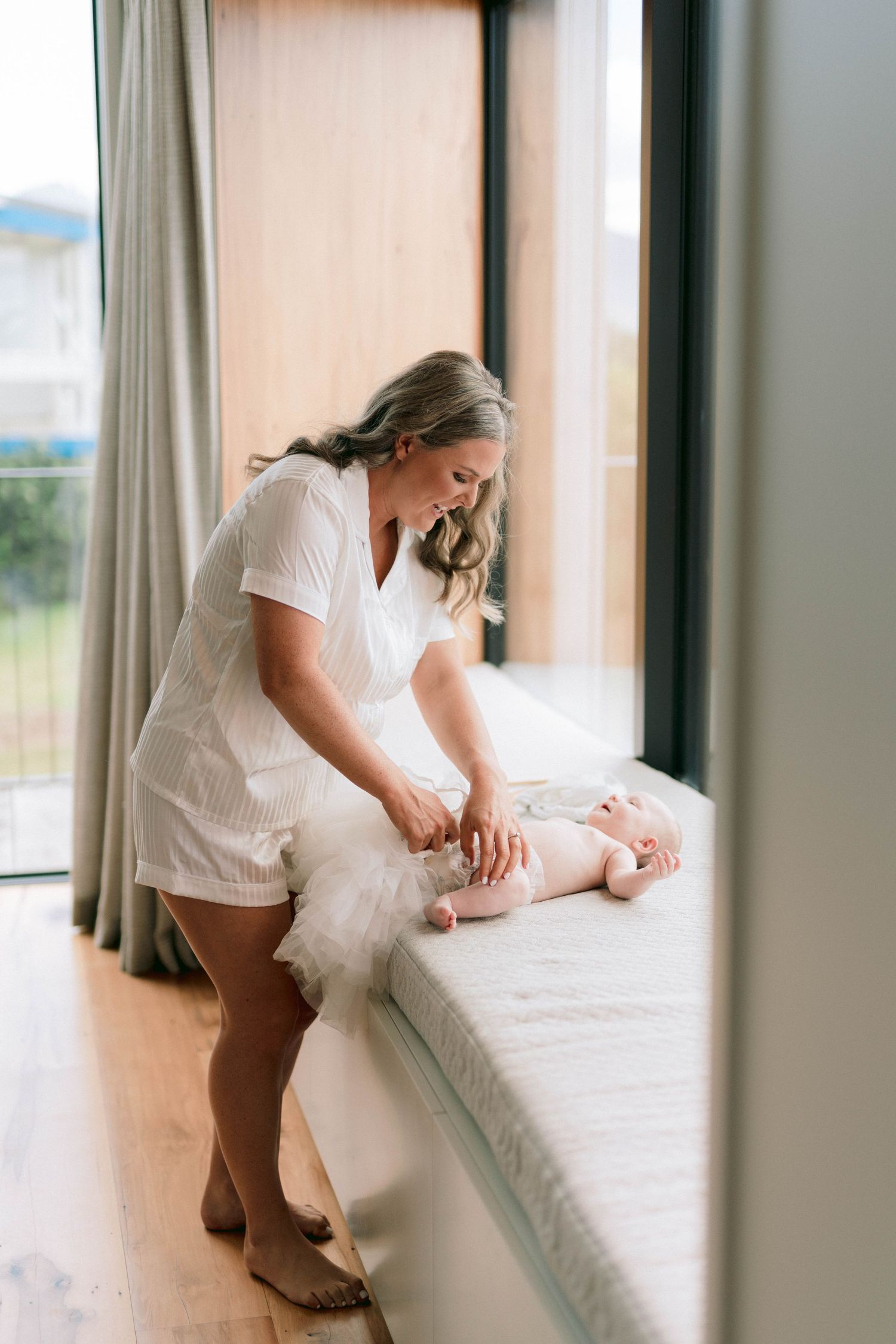 Bride in white shorts gets ready in sunlit room while arranging something on white bedding.