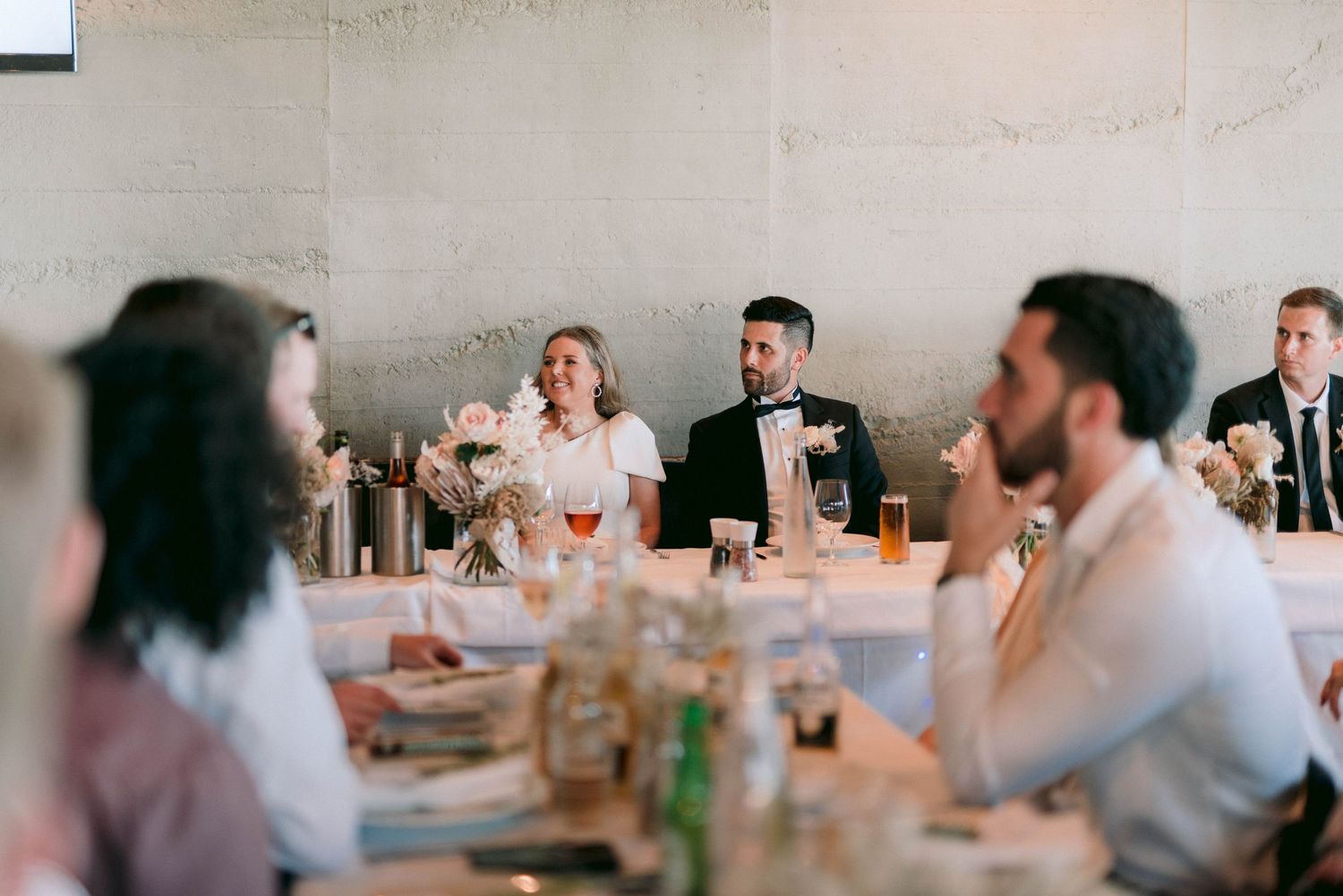 Wedding reception guests gathered at long tables in elegant indoor venue with ambient lighting.