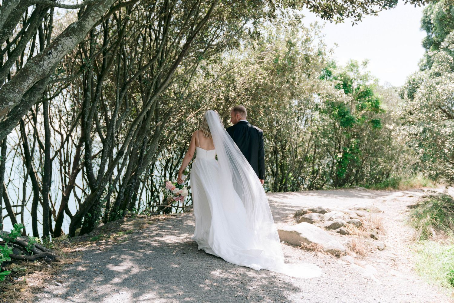A flowing white wedding dress trails gracefully along a forest path bordered by trees.