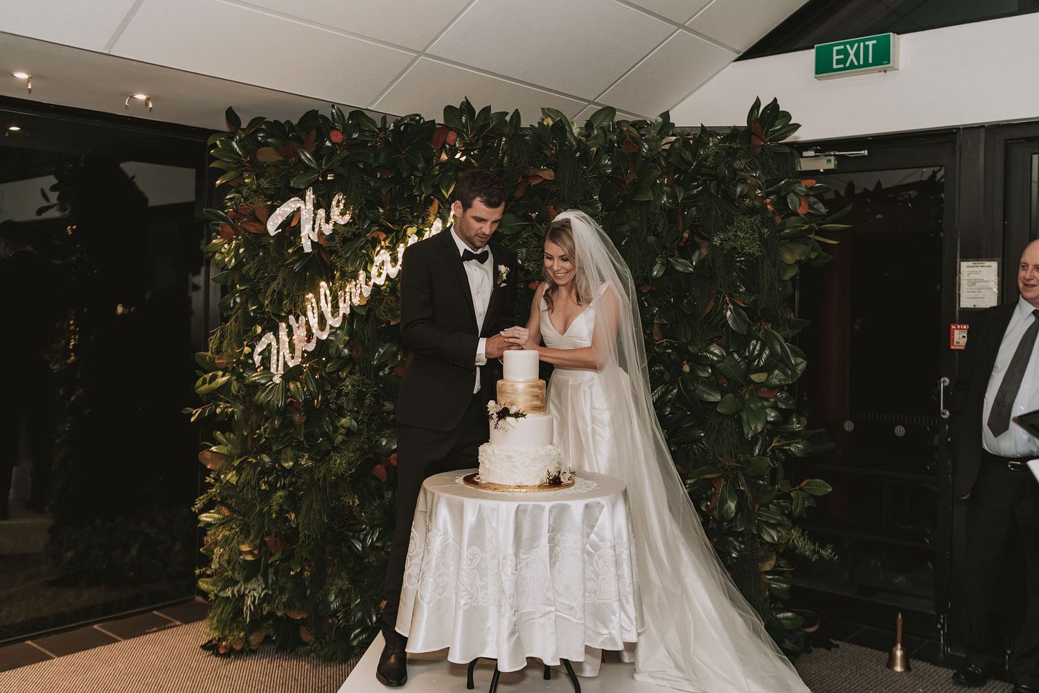 Bride and groom cut their elegant white wedding cake together in front of a lush greenery wall with neon signage.