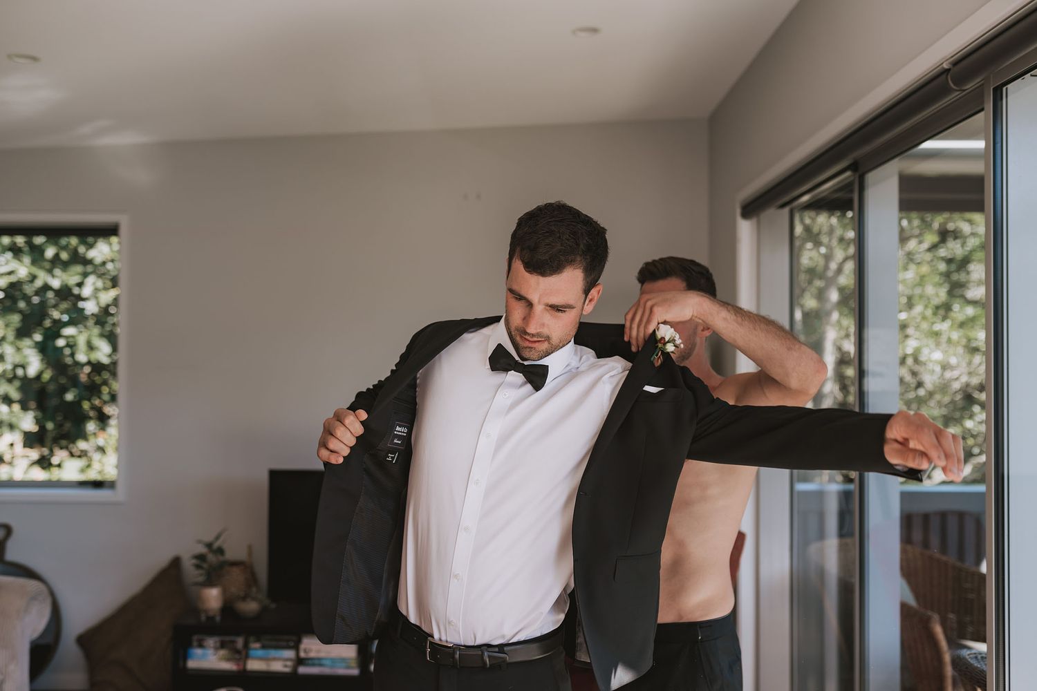 A person in formal attire puts on a black suit jacket while getting dressed in a sunlit room.