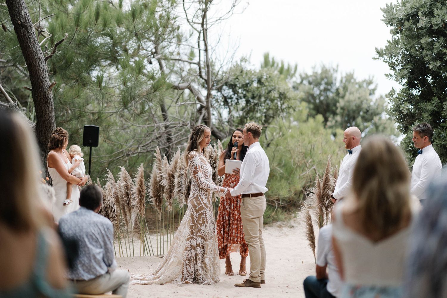 Intimate outdoor wedding ceremony in a natural setting with pampas grass decorations and guests watching the couple.