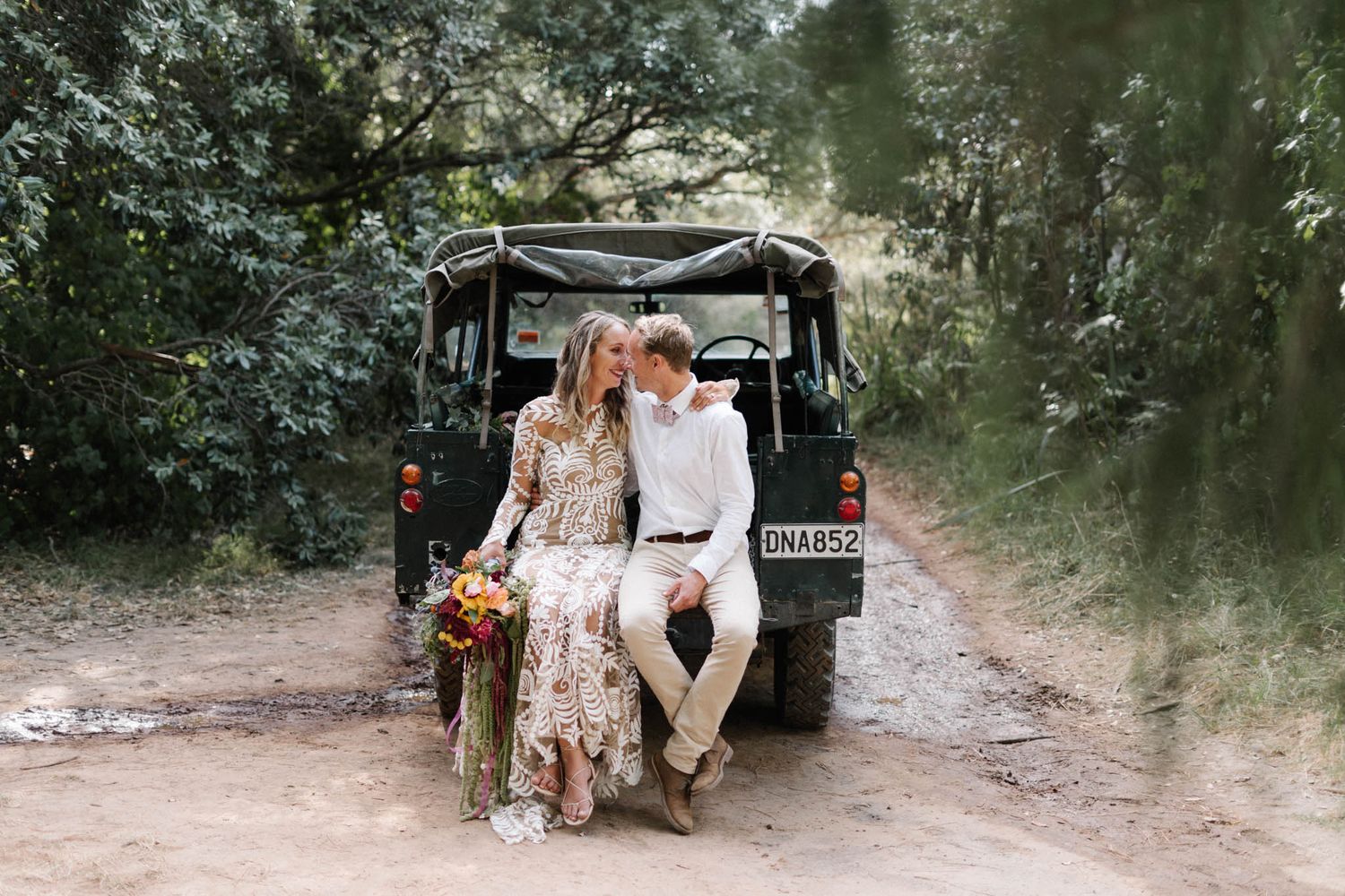 Two people sit together at back of vintage black Land Rover Defender on forest dirt road with floral bouquet.