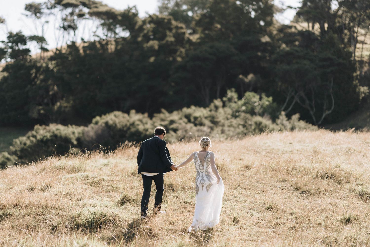 A couple walking together in an open golden field with trees in the background.