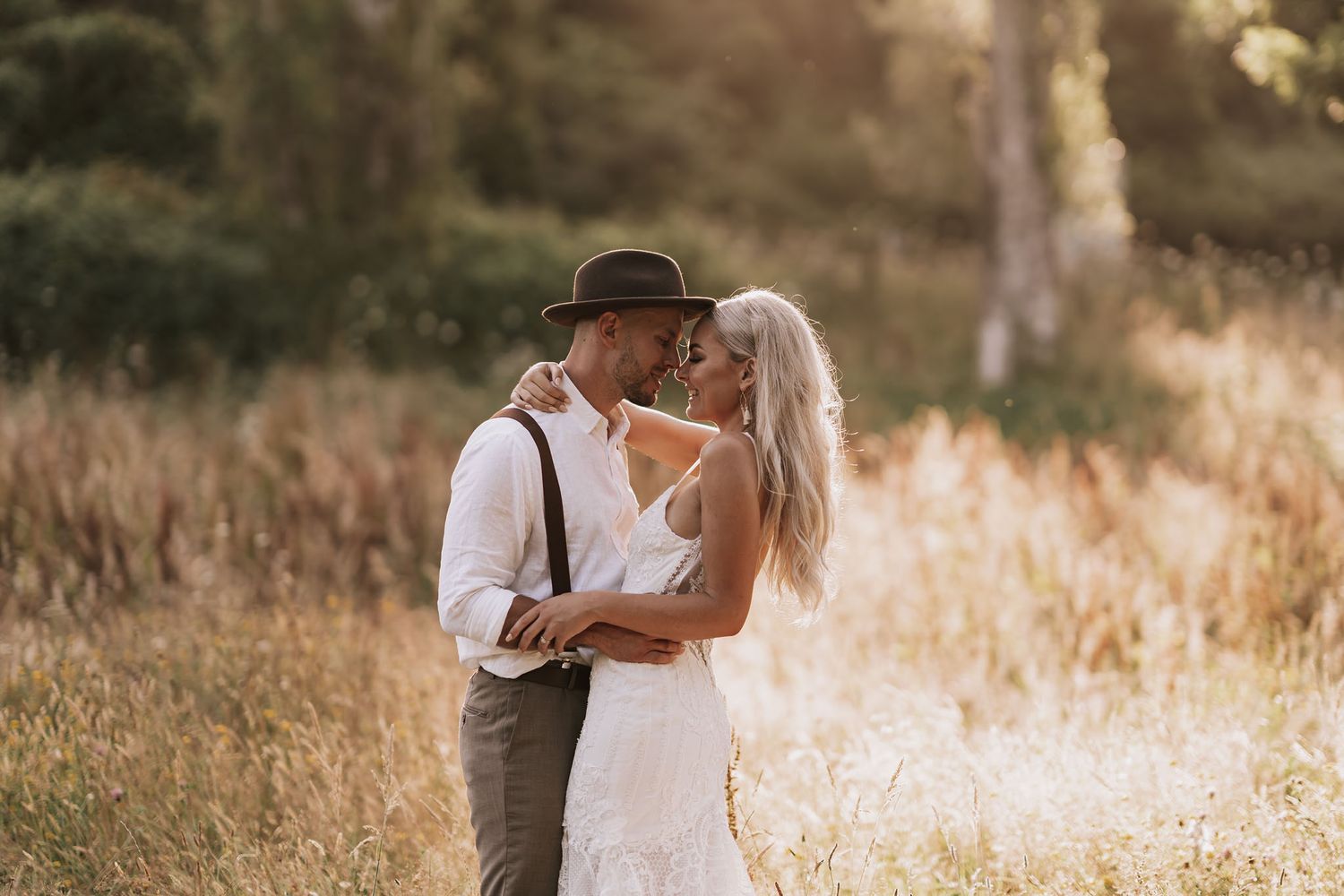 A romantic couple in wedding attire embrace in a sunlit grassy field at golden hour.