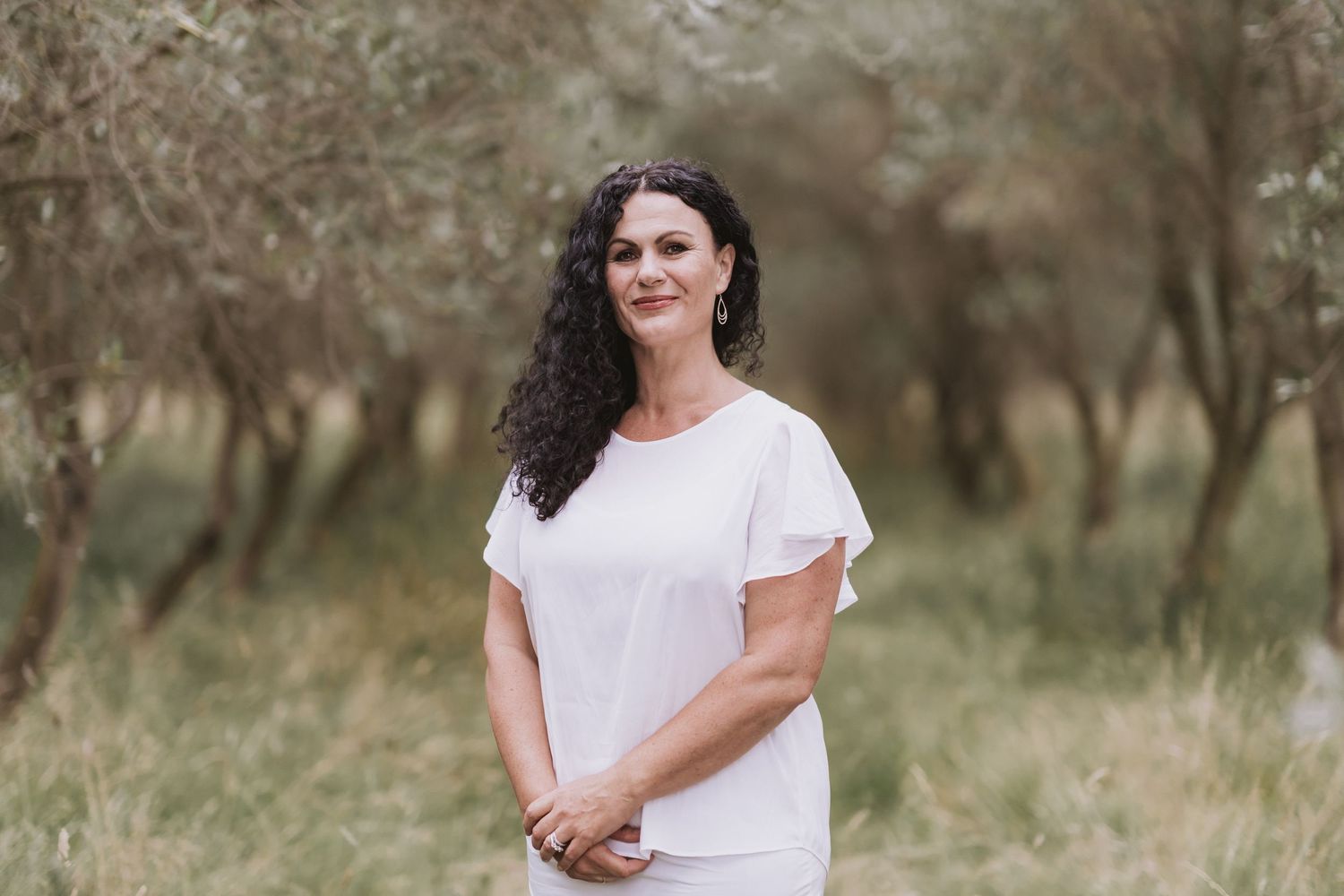 A person in a white short-sleeved top stands in a serene outdoor field with blurred trees in the background.