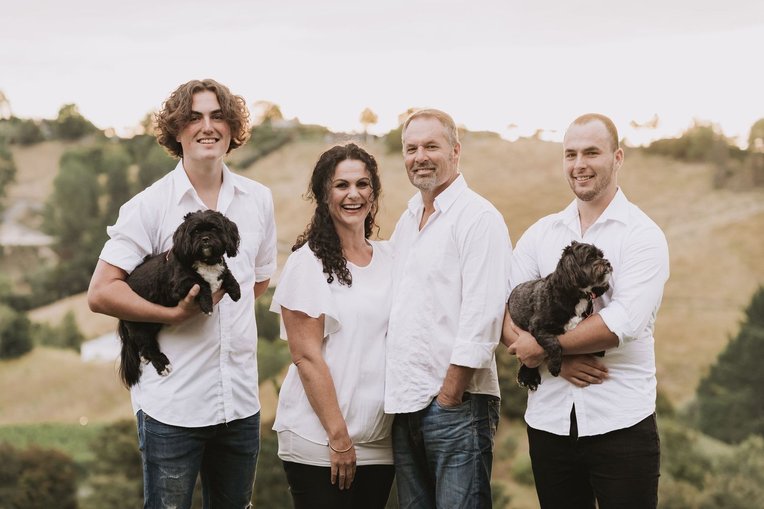 Family portrait in white shirts on a hillside with two adorable black puppies held by people on each end.