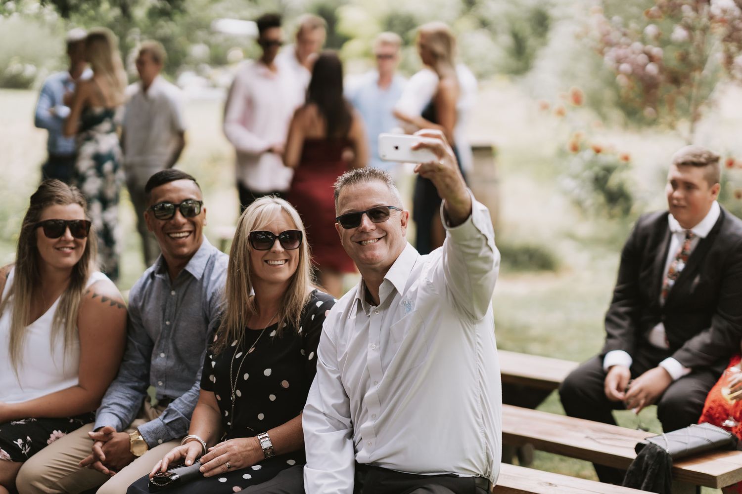 A group of well-dressed guests sit on outdoor benches at a casual outdoor gathering in a garden setting.