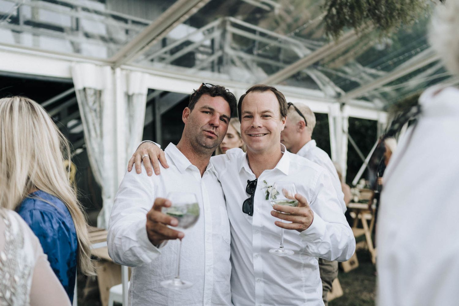 Two people in white clothing smiling and holding drinks at an outdoor party venue.