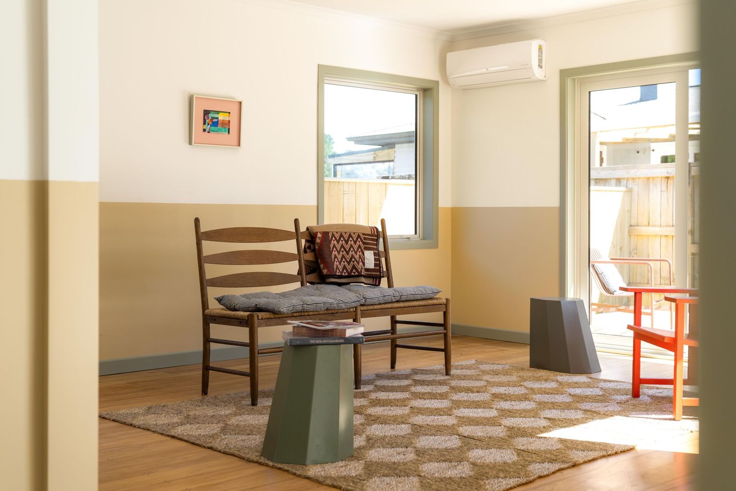 Vintage wooden bench and green pedestal table sit in a minimalist room with two-tone walls and patterned floor tiles.