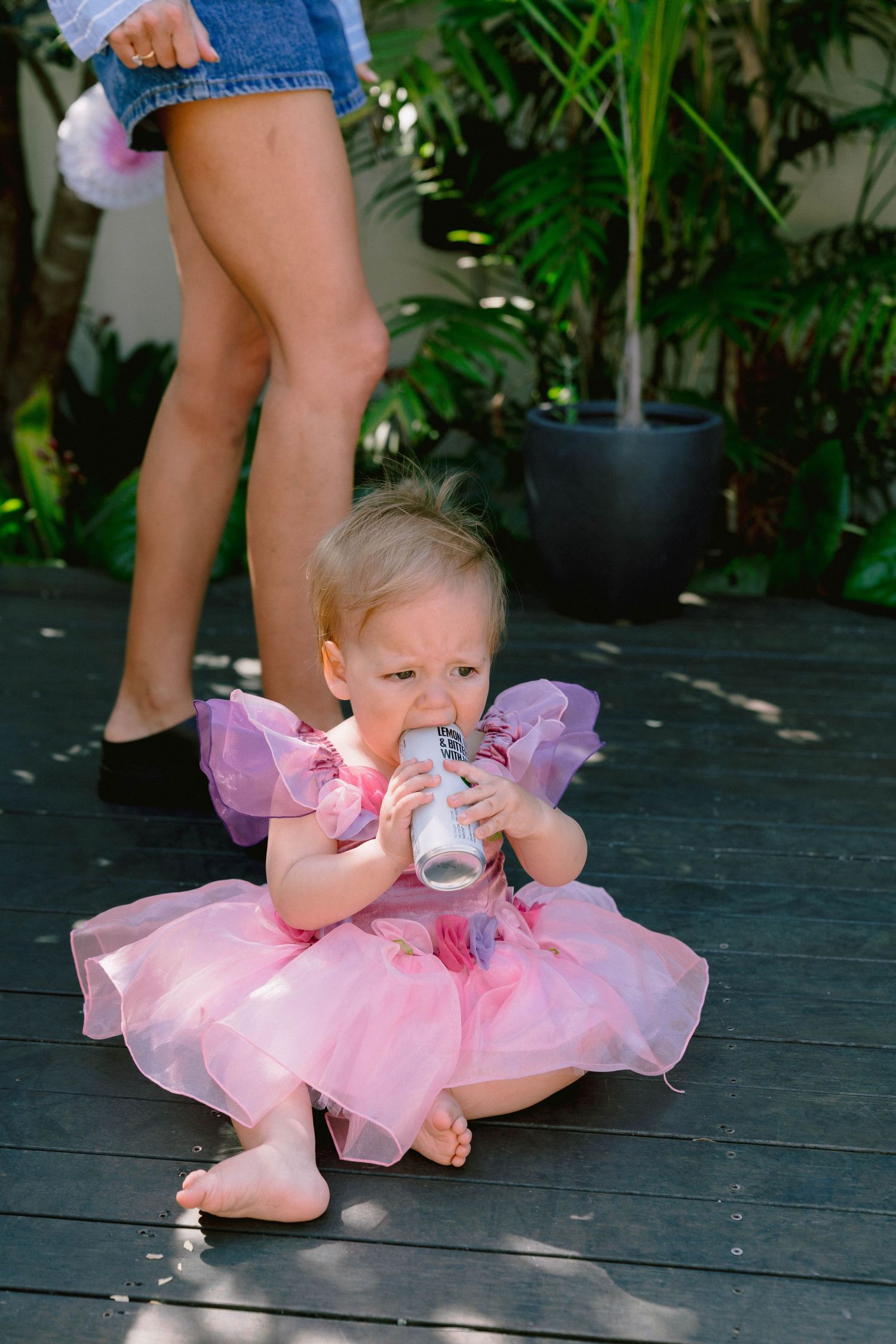 A baby in a pink tutu dress lying on a dark wooden deck while holding a small bottle.