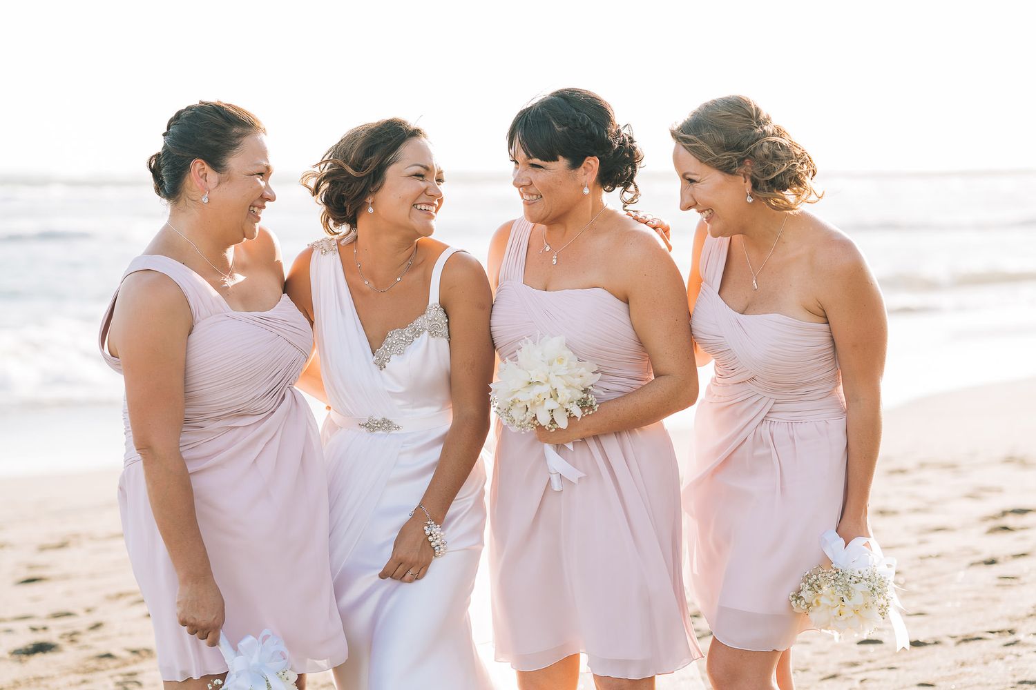 Bridesmaids in light pink dresses laughing together on a sunny beach.