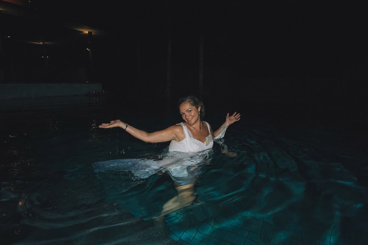 Person in white dress floating in illuminated swimming pool at night with arms outstretched.