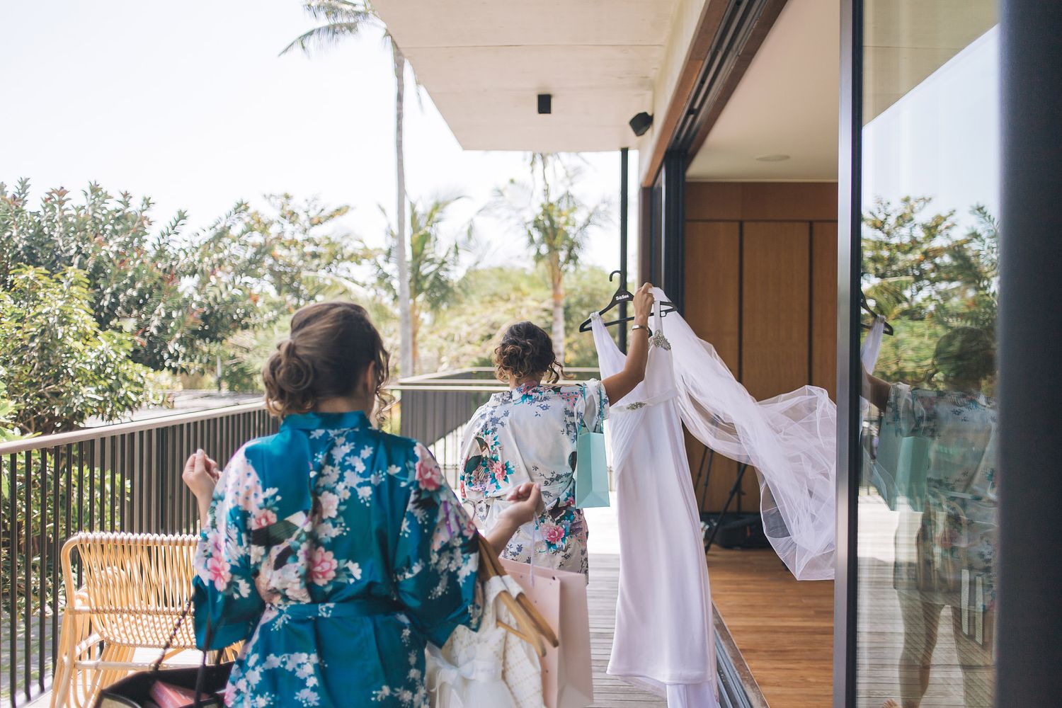 Bridesmaids help with wedding dress preparations in a bright room with natural lighting and white curtains.