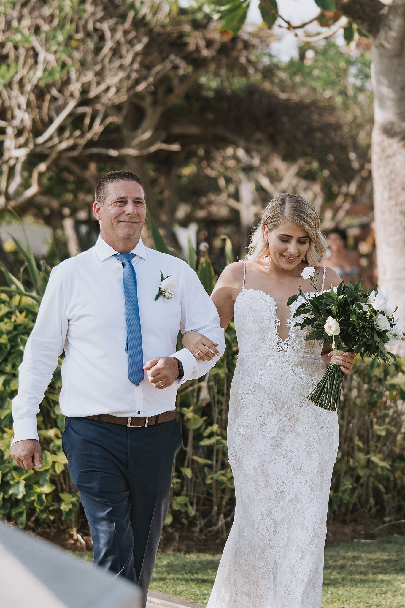 Man in blue tie and white shirt walking with bride holding white flower bouquet.