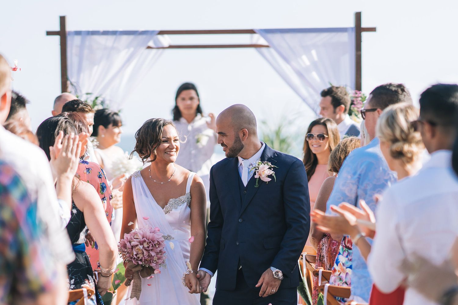 Wedding couple exchanges vows under a white fabric arch during an outdoor ceremony.