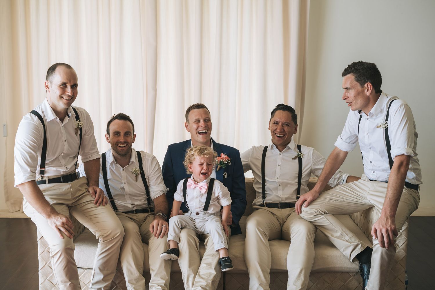 A group wearing matching khaki pants and suspenders sit casually together on a couch during a formal event.