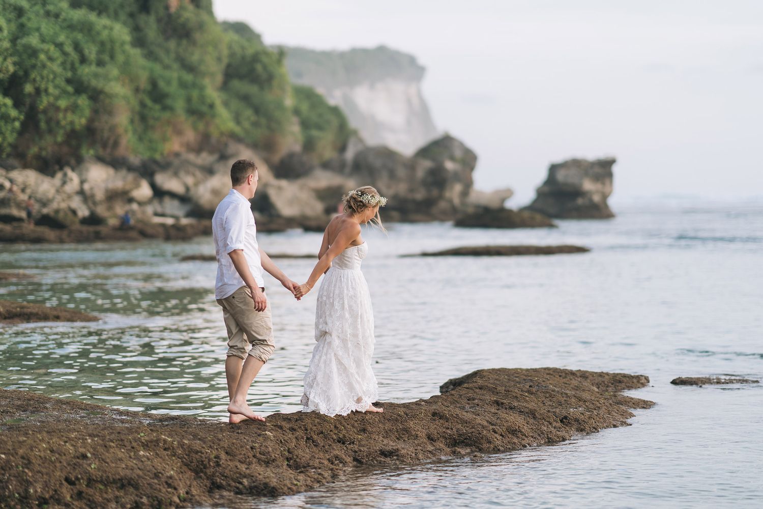 Couple stands hand in hand on rocky coastline with cliffs in the background.