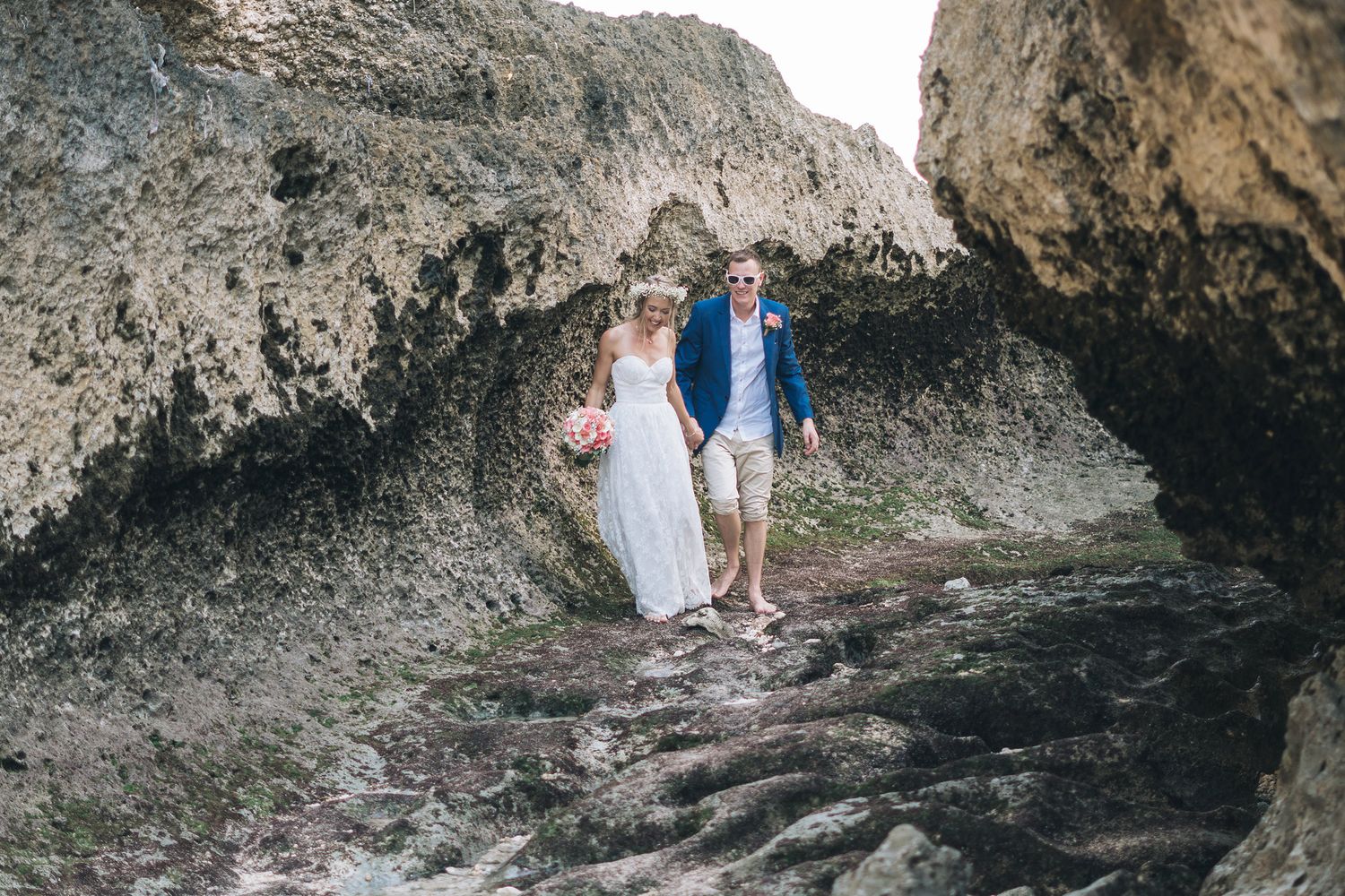 Couple in wedding attire standing between rocky cliffs on a beach.