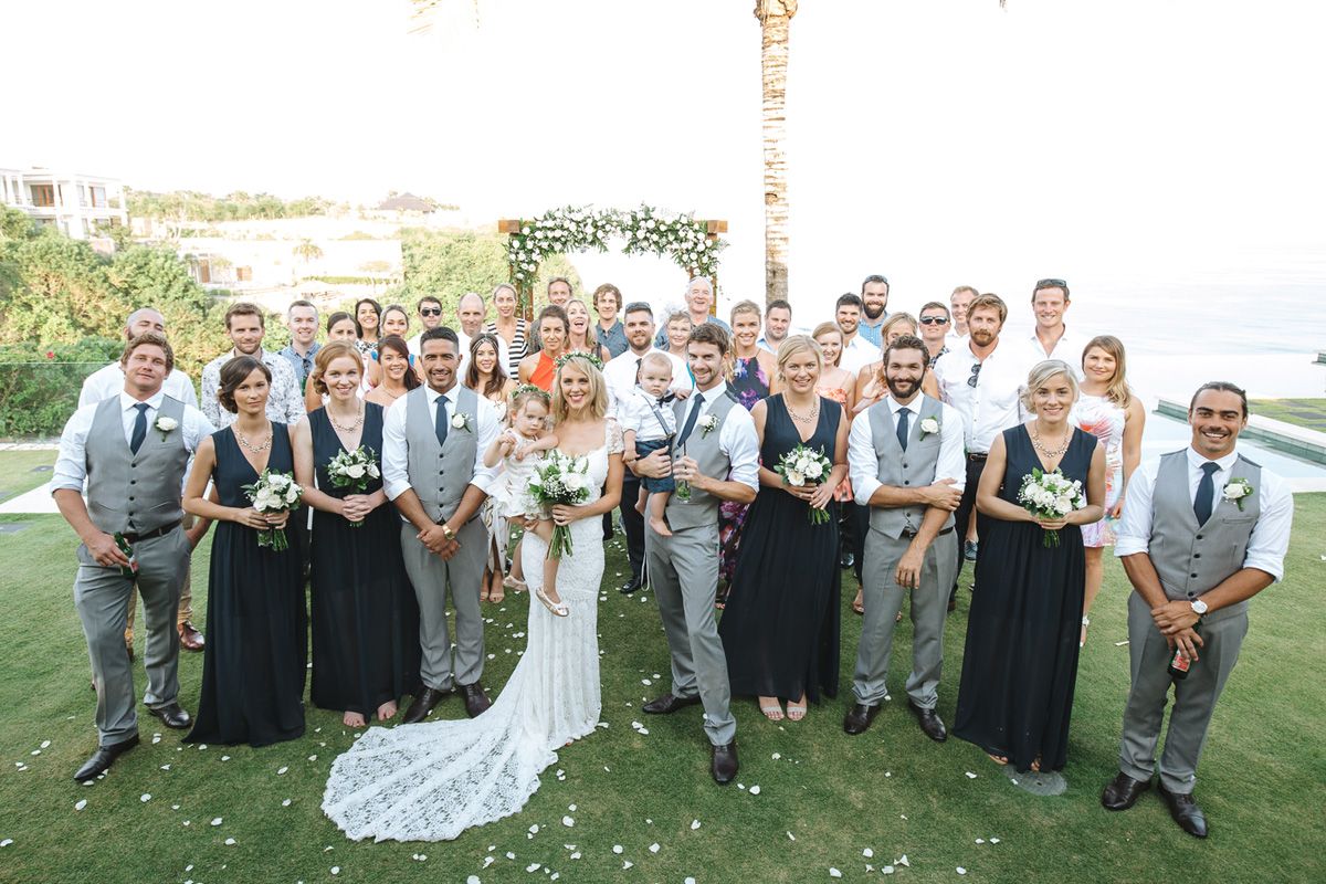 Large wedding party in gray and black attire poses together on a sunny lawn with ocean views.