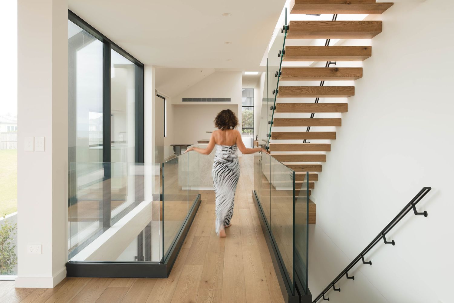 Person ascending modern floating staircase with glass railings and hardwood floors in bright contemporary home interior.