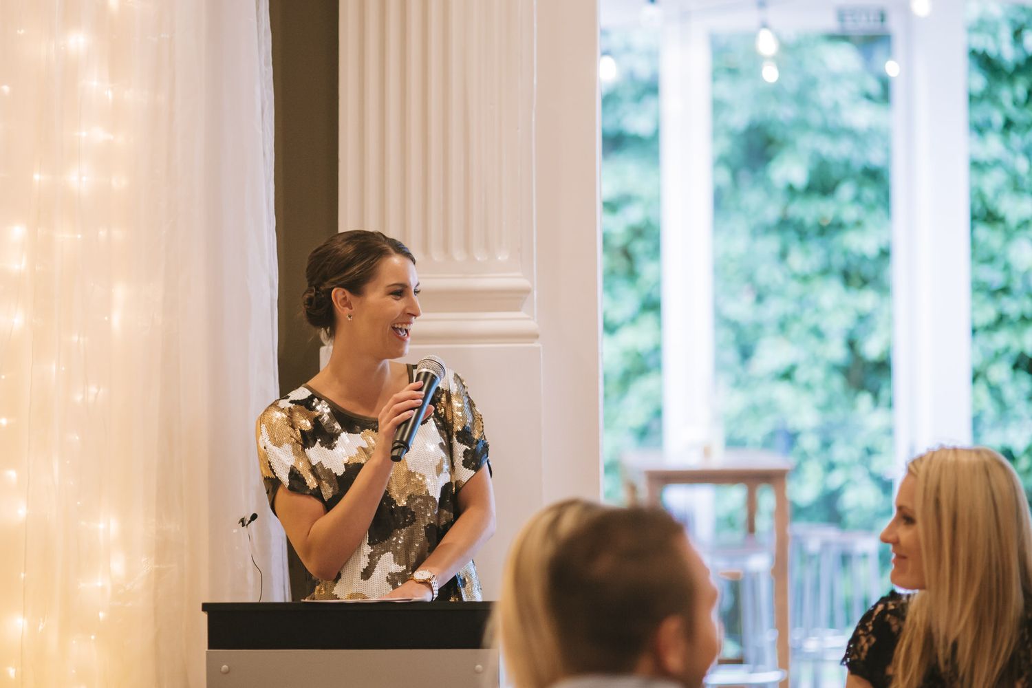 A speaker at a podium delivers a presentation in front of a window with string lights and white columns.