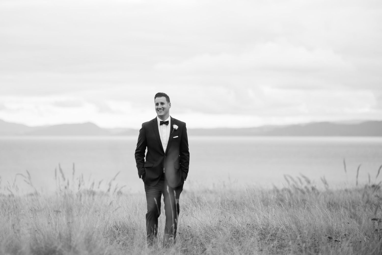 A groom in a black suit and tie stands alone in windswept tall grass against a misty mountainous landscape.