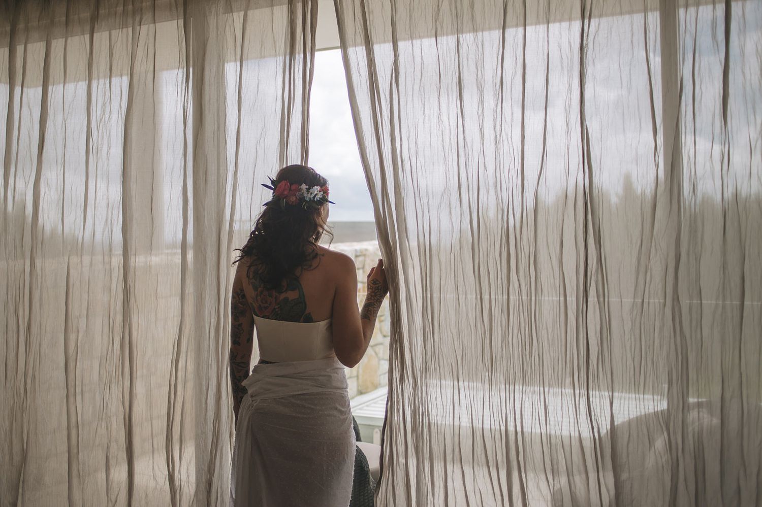 A bride in a white wedding dress peers through sheer curtains at a window on her wedding day, wearing a flower crown.