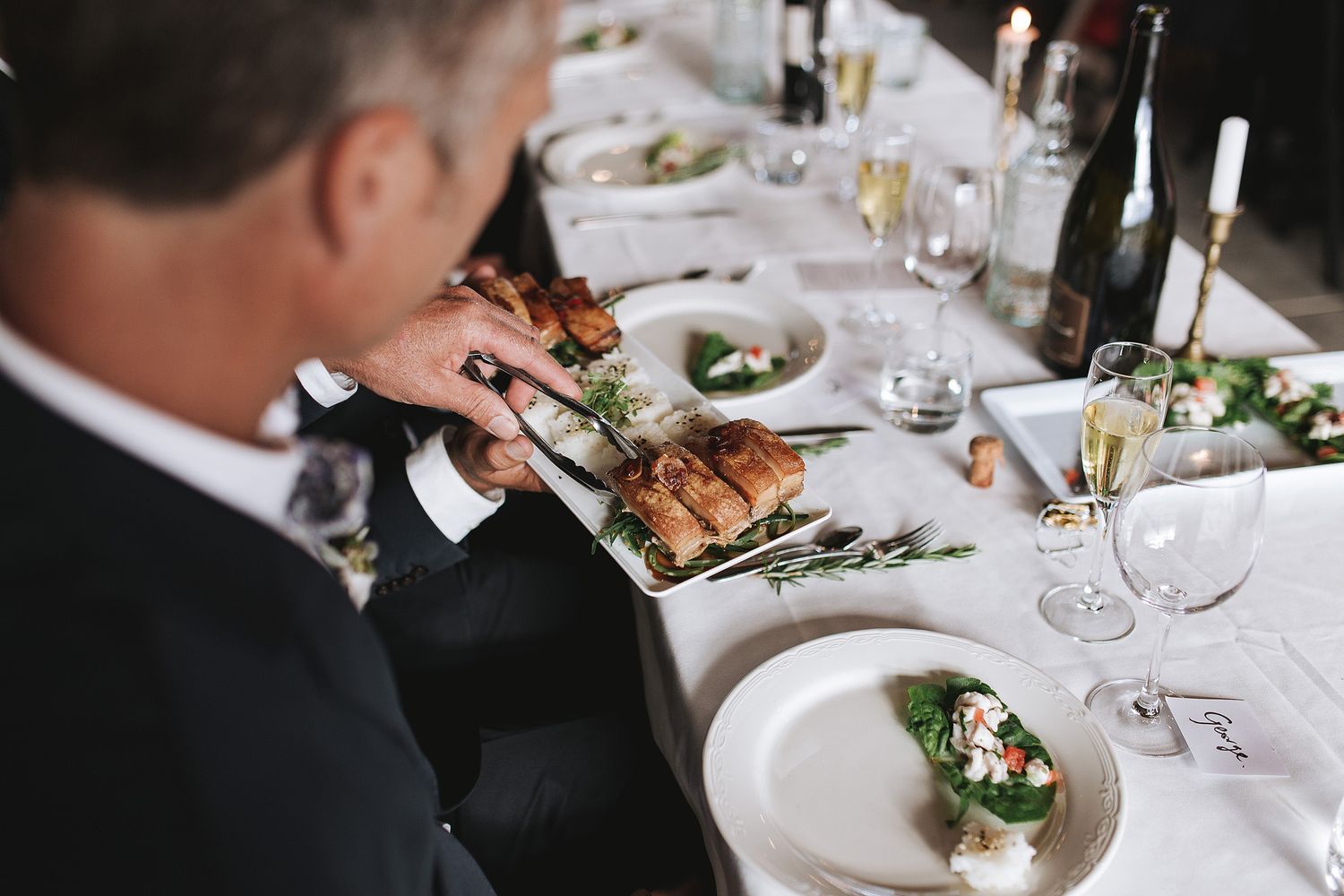 Elegant table setting at formal dinner party with white plates and person in black suit reaching for food.