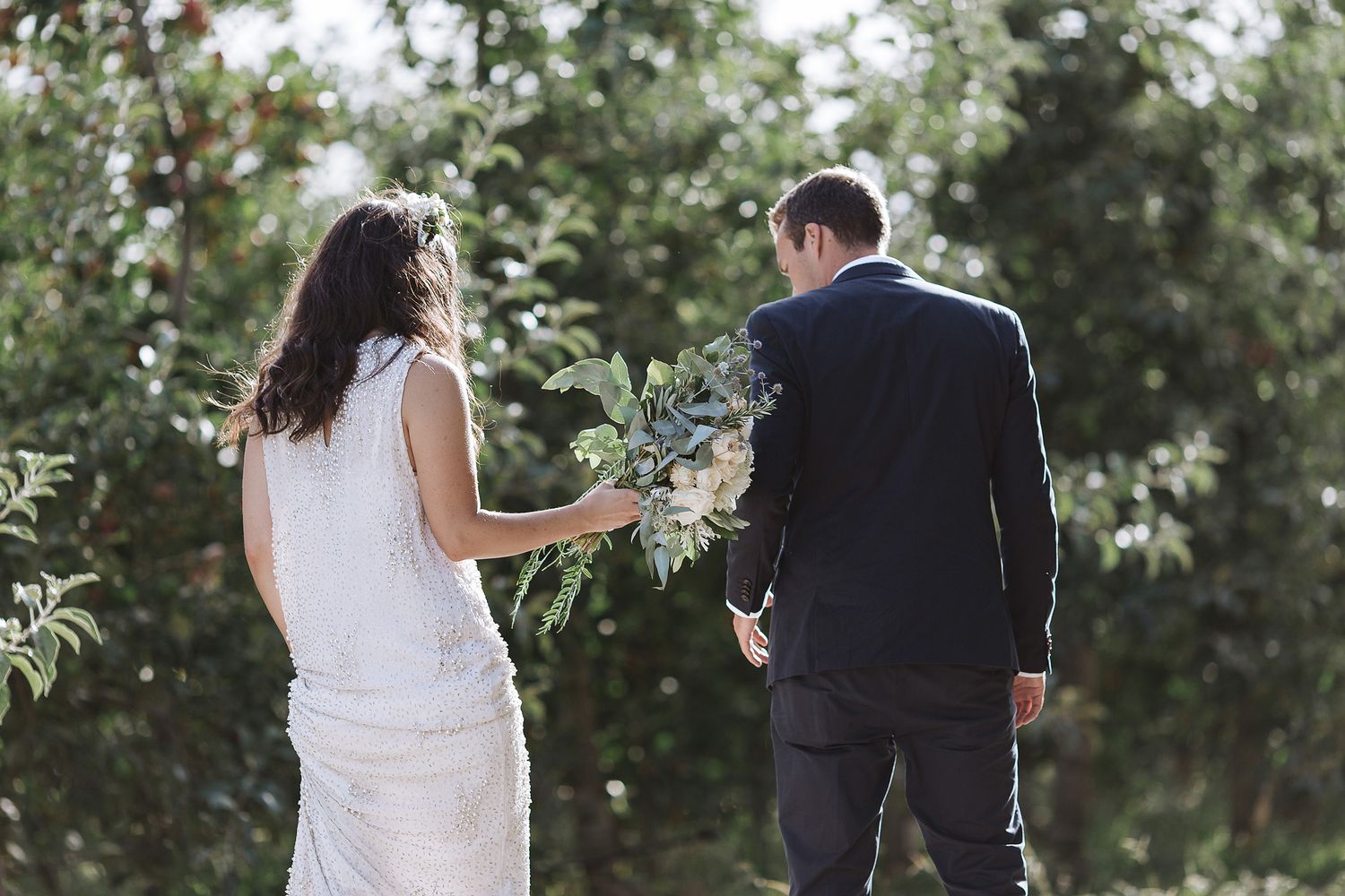 A couple in wedding attire walks together outdoors with trees and greenery in the background.