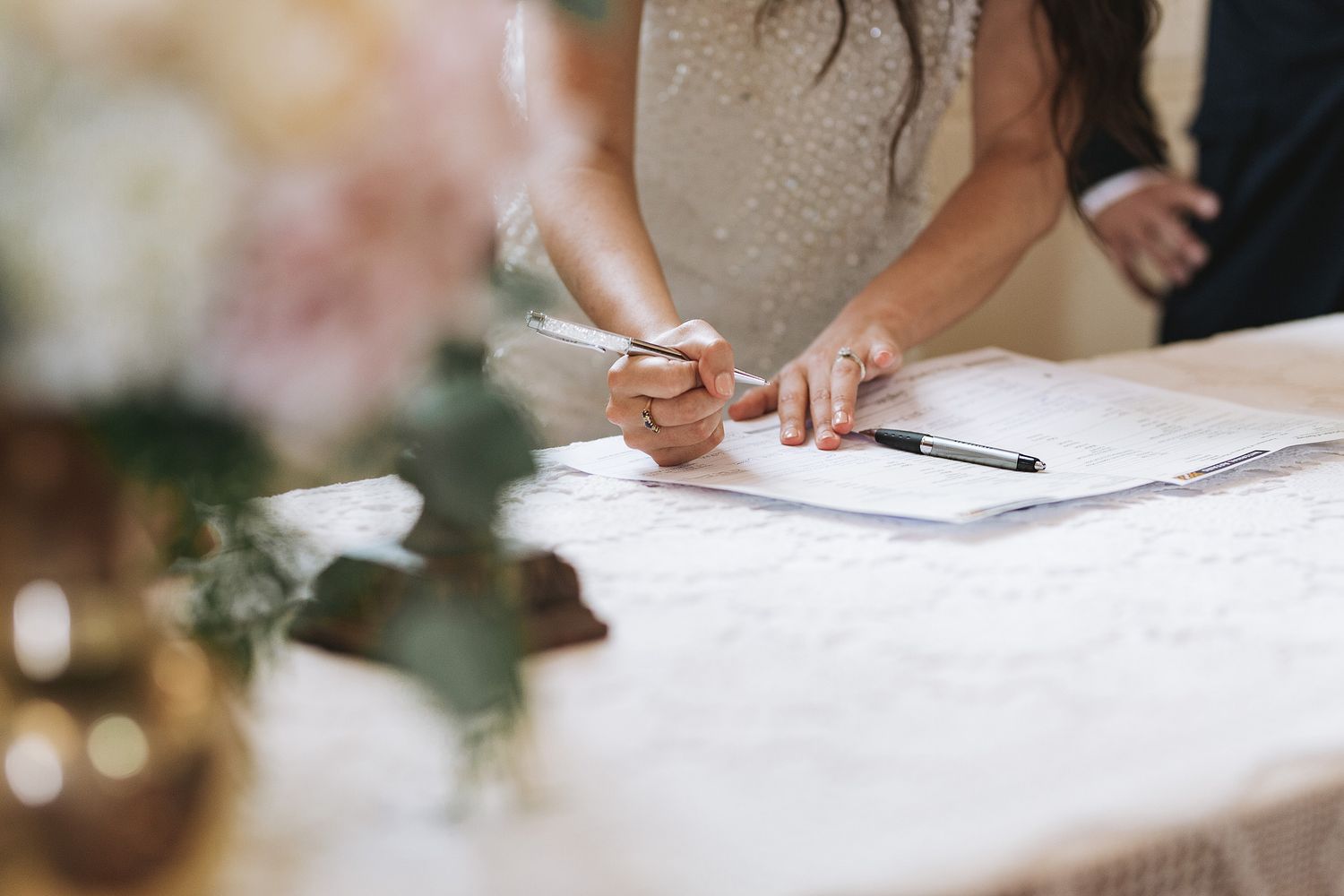 Someone signs a document at an event with decorative floral arrangements in the background.