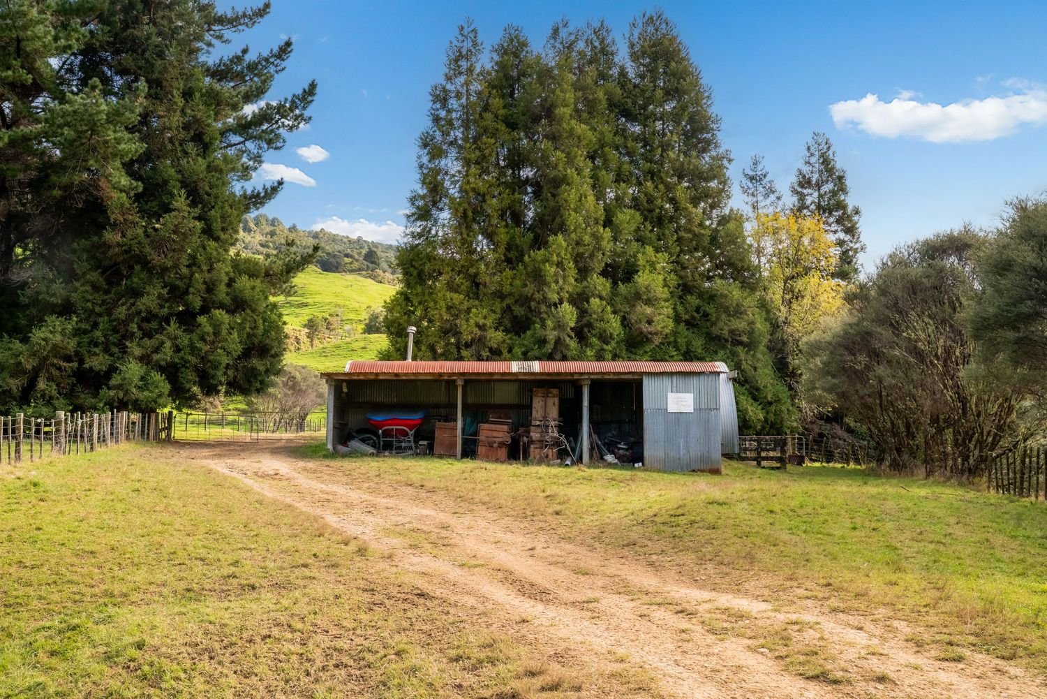 Small farm shed with red panels situated on grassy hillside with tall evergreen trees.