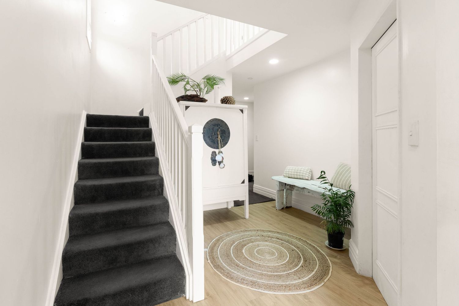 Modern white hallway featuring dark carpeted stairs, circular rug, and decorative wall mirror.
