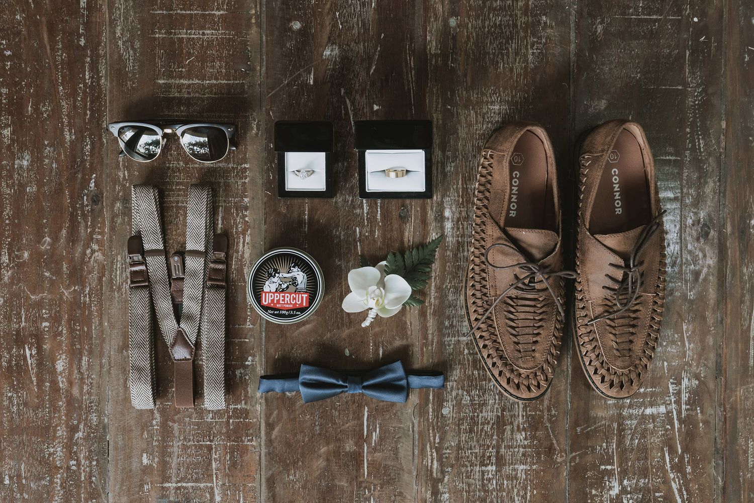 Wedding accessories including bow tie, suspenders, shoes, and glasses arranged on wooden surface.