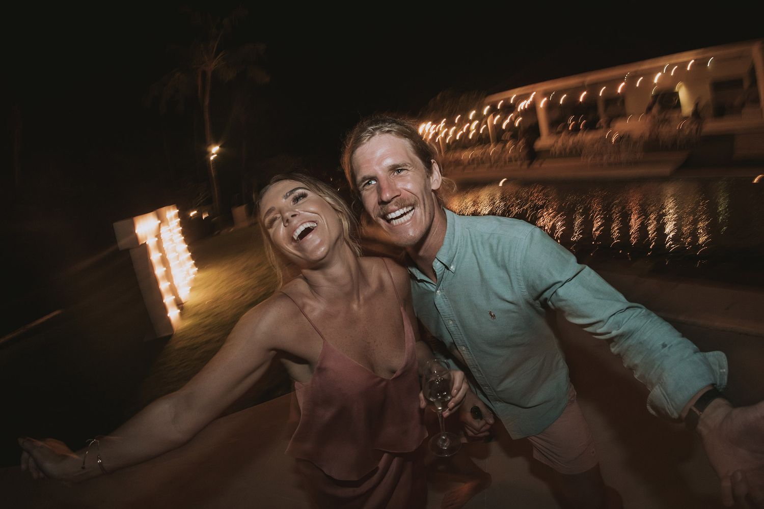 Two people laugh together at night with decorative lights visible in the background at an outdoor venue.