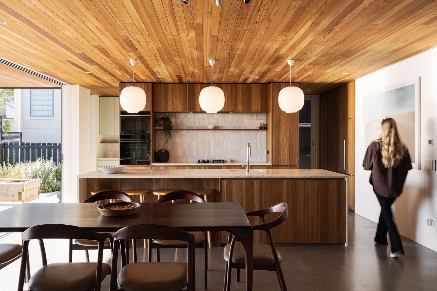 Modern open-plan kitchen with warm wood ceiling, island bench, globe pendant lights, and dining area in contemporary home.