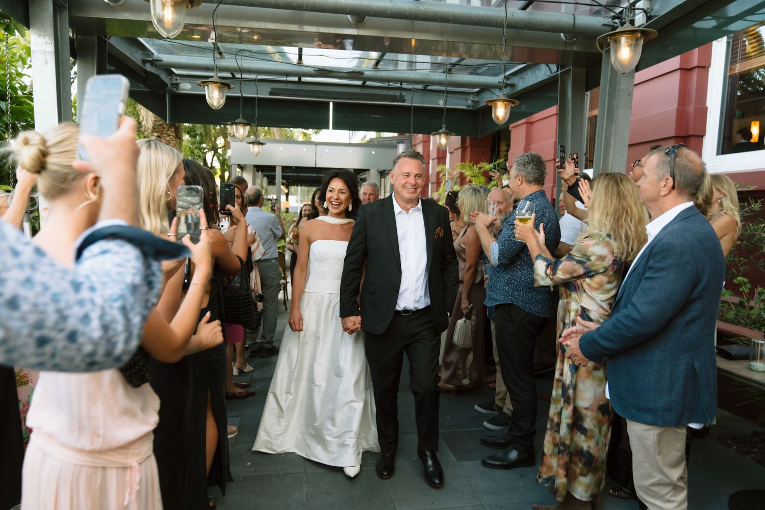 Bride and groom walk through cheering guests at outdoor wedding reception while attendees photograph the celebration. Bryony Alexander.