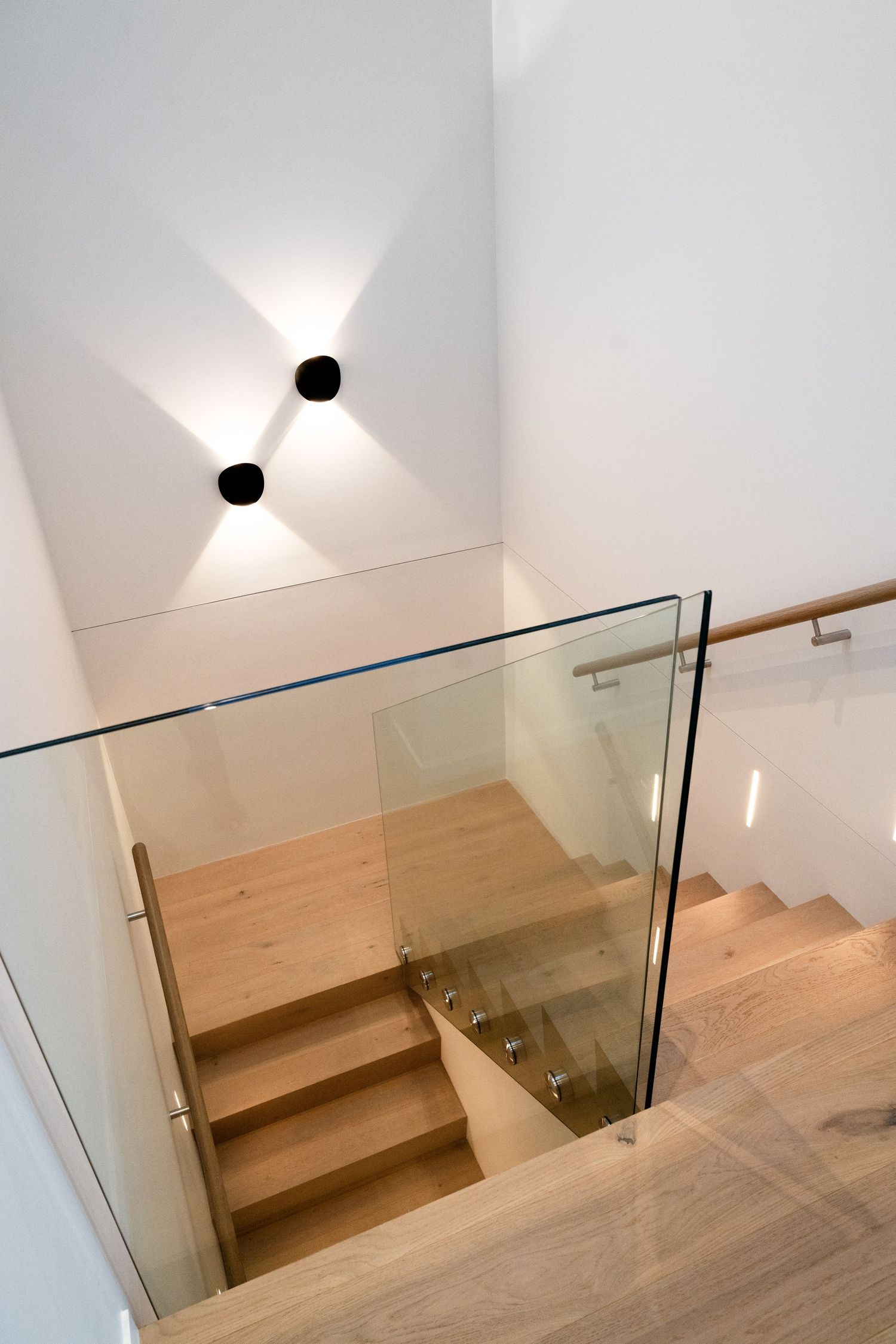Modern wooden staircase with glass railing and minimalist wall sconce lighting creates an elegant interior design statement.