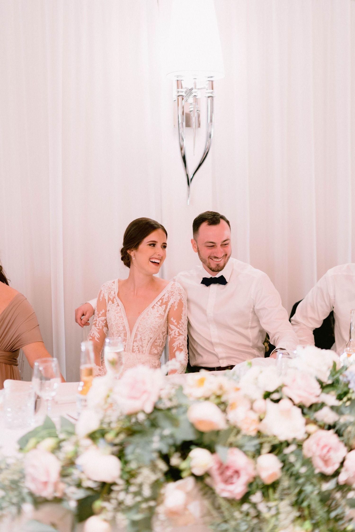 Wedding couple smiles at their reception table decorated with pink floral centerpieces.